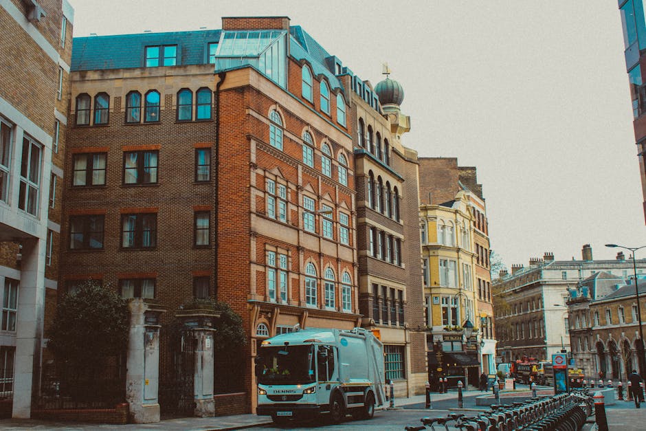 A street scene in an urban area showing a white removal van parked close to a row of multi-storey buildings with a variety of architectural styles, including brick and stucco facades. The van is situa