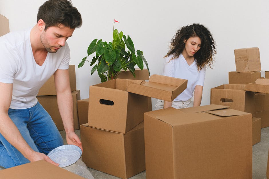 A man and a woman stand indoors in a room with sloped ceiling, each holding large cardboard boxes filled with household items, positioned next to a wooden bed frame. The man has dark hair, a beard, an